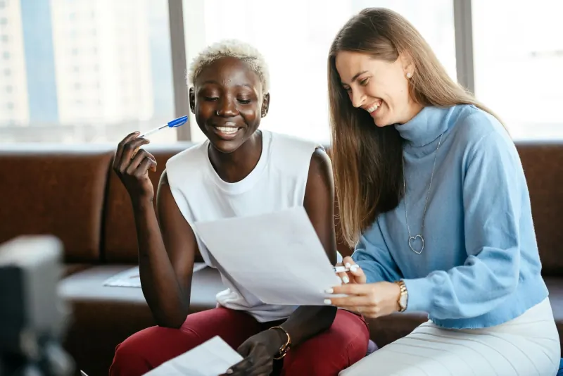 Two women collaborating on paperwork at a sunlit window, smiling